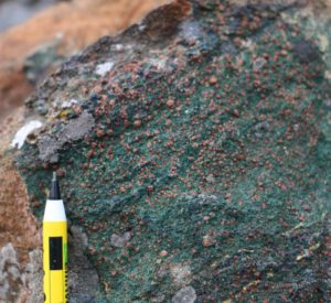 A close-up photo of a green rock with prominent red garnets (shaped like small soccer balls). A pencil tip provides a sense of scale.