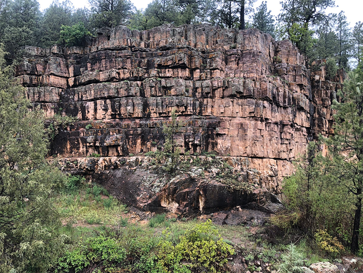 A photo of a big cliff-like outcrop, surrounded by forest. At the bottom are rounded knobs of granite (labeled 1.7 Ga) and above that are regular blocky layers of quartzite (labeled Devonian).