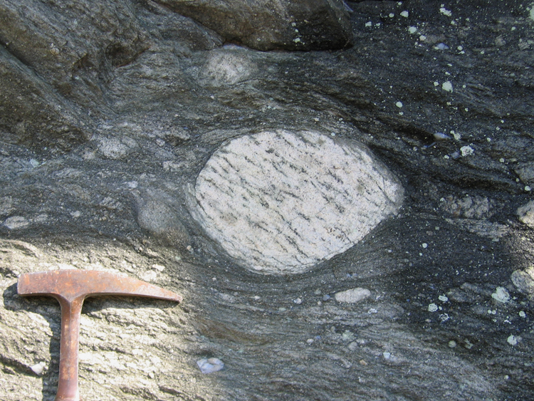 Photograph showing a prominent outsized clast of gneiss in metadiamictite. Foliation and bedding are both horizontal, wrapping around the clast. A rock hammer provides a sense of scale.