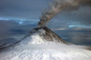 Photograph of a volcano erupting, with a plume of ash (eruption column) drifting off to the right. There is snow on the volcano.