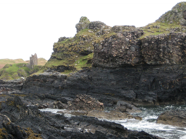 Photograph showing a seaside cliff with very coarse conglomerate at the top and fine-grained metamorphic rocks at the bottom. There is an old castle ruin in the misty distance. A big boulder of the conglomerate has fallen down.