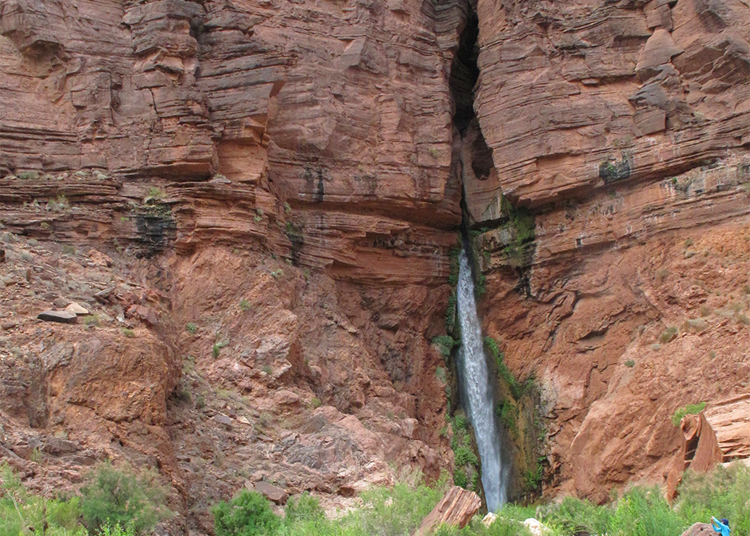 A photograph showing a waterfall emerging from a slot canyon right at the contact between thin upper layers of sandstone (horizontal) and a lower cliff of link granite and gray schist.