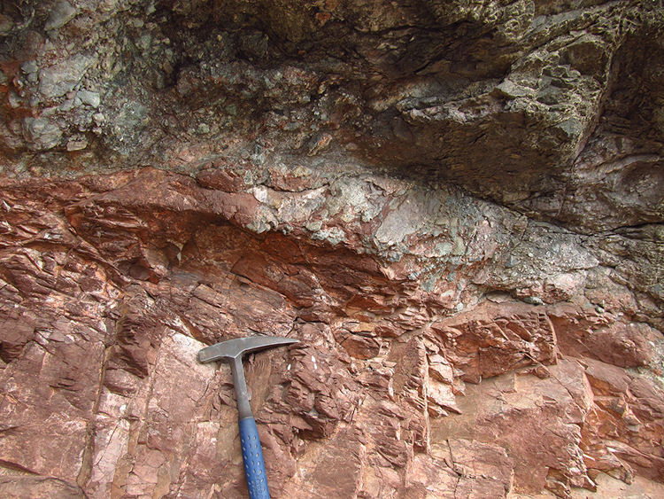 A photo showing the horizontal contact between two rock units: a lower fine grained red unit, and an upper greenish chunky unit. A hammer provides a sense of scale.