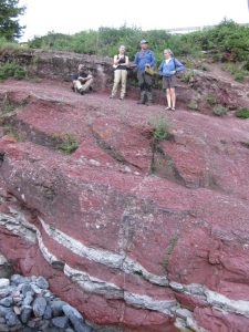Photograph showing a cliff of red and white layered rock, about 9 m tall. The layers dip gently to the right. Four geologists stand at the top. Most of the cliff is red, but htere are two prominent white layers at the bottom, each about half a meter thick. Some dark colored boulders (a streambed) are at the bottom of the cliff.