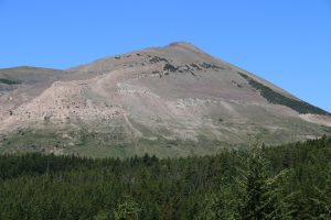 Photograph showing a mountain with outcropping layers of green, white, and purple rocks. The layers make a