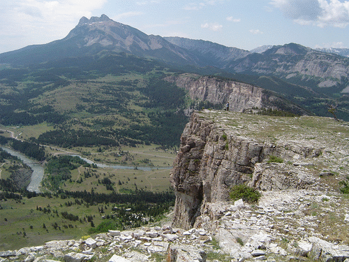 Animated GIF of an annotated photograph showing a view to the south across the landscape of the Sawtooth Range at Sun River Canyon. East is to the left and west is to the right. The Sun River can be seen in the middle distance at left center. In the foreground are cliffs of white Mississippian carbonate, and these are repeated in the middle and far distance, separated by the traces of faults from gentle vegetated slopes underlain by Cretaceous siliciclastics.