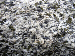 Photograph showing an outcrop of granite, measuring about 20 cm wide by 15 cm tall. White feldspar and blue quartz are the most prominent minerals. A few patches of brown/green lichen are growing on the rock's surface.