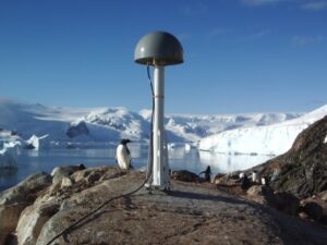 Photograph of a GPS antenna in Antarctica. The antenna looks like a stick with a fat dome on top, kind of like a toadstool. It's anchored to rock, and in the background is a glacier, some mountains, and a curious penguin.