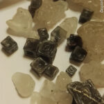 Photograph showing halite gravel from the Dead Sea: a mix of light colored blobby looking bits and very striking dark colored "hopper" cubes. A pen serves as a sense of scale.