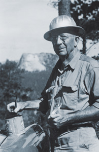 Black and white photograph of a man standing, wearing a baggy field shirt and a pith helmet. He is holding a bottle in one hand. There are mountains in the background.
