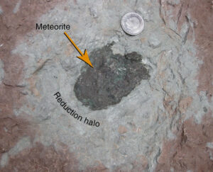 Photograph showing a big lumpy black meteorite sitting in red limestone, with a light gray zone of reduced chemistry surrounding the meteorite (a "reduction halo"). A coin serves as a sense of scale.