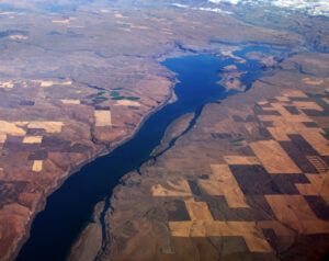 Aerial photograph over a long lake that partially fills a narrow long valley with step-like cliffs walling it in. A grid of farm fields covers the neighboring upland plateau. Snowcapped mountains loom in the distance.