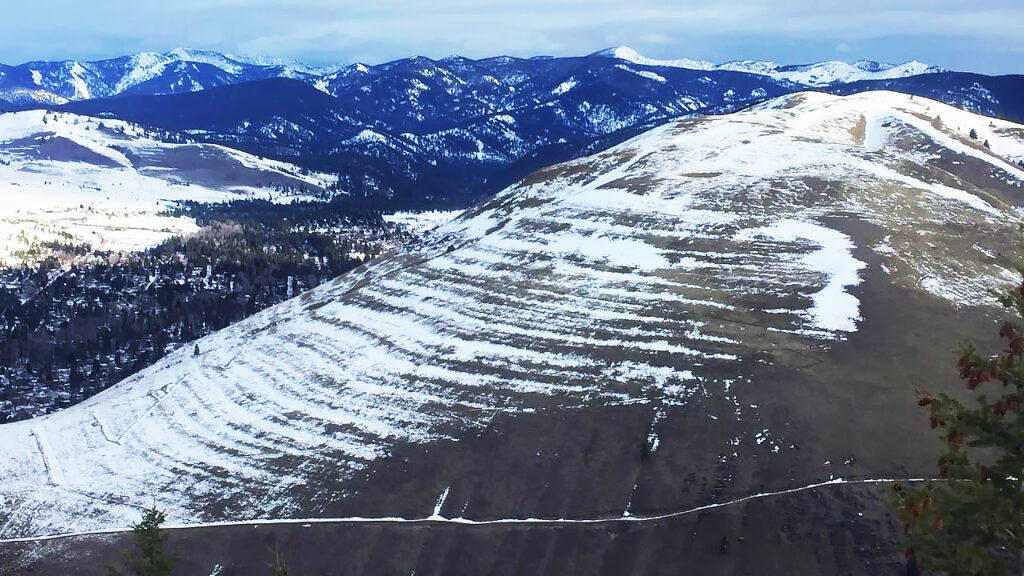 Aerial oblique photo of a partially snow-covered hillside, with ~40 horizontal "contour lines" wrapping around the hillside. 