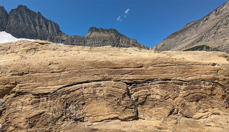 A photograph of a mountainside, with a blue sky above and some snow. Most prominent is in the foreground there are a series of four stromatolites - laminated mounds in the rock, viewed in cross-section.