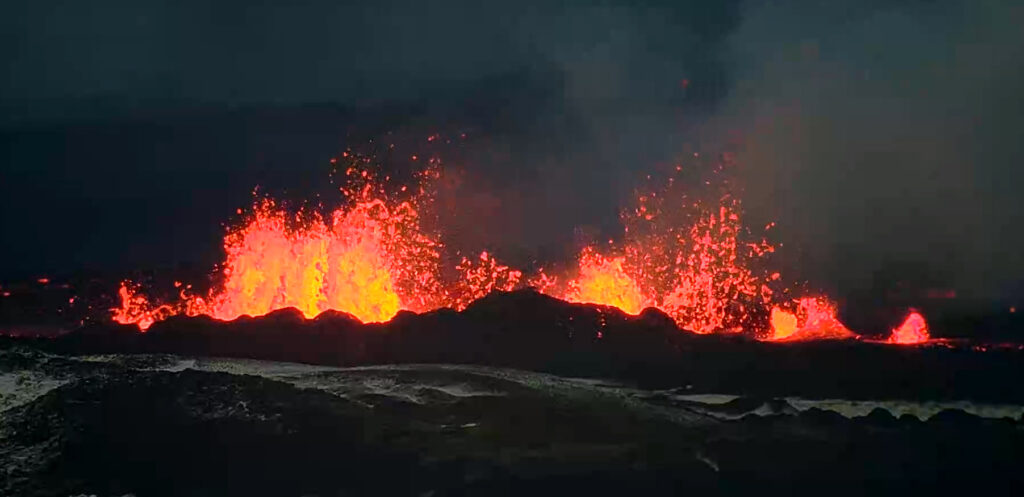 Photo showing incandescent lava fountaining upward from a crack that runs in a line left to right across the field of view. In front of the orange splattery lava fountains are black lumpy rock fields, dusted with white snow. Steam rises to the rear of the lava fountains and drifts away to the right.